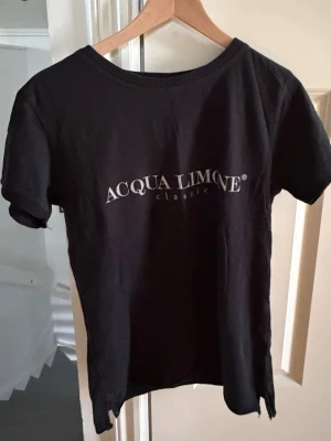 Svart Acqua Limone t-shirt - Snygg svart t-shirt från Acqua Limone med klassisk logga framtill och stor gul citron-tryck på ryggen. T-shirten har rund halsringning och korta ärmar. Tillverkad i mjuk bomull som känns skön mot huden. Perfekt för en avslappnad och trendig stil.