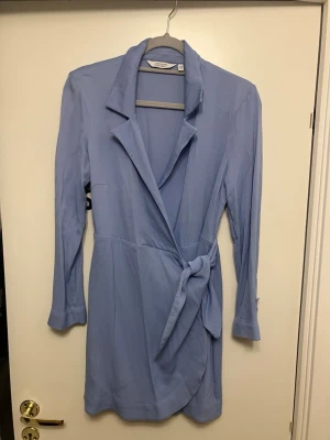 Ljusblå omlottklänning & Other Stories - Snygg ljusblå omlottklänning från & Other Stories med lång ärm och knyt i midjan. Klänningen har klassisk krage och en avslappnad passform som ger en chic look. Perfekt för dig som vill ha något stilrent och trendigt. Snöret man knyter med är lite trasig, se bild! Annars jätte fin