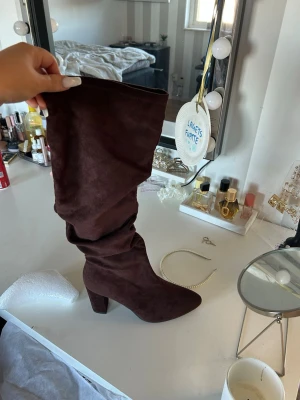 Bruna höga stövlar i mockaimitation - Säljer ett par snygga bruna stövlar med hög klack och slouchy skaft i mjuk mockaimitation. Perfekta att styla till kjol eller jeans för en trendig look. Stövlarna har spetsig tå och ger en cool vibe till outfiten.