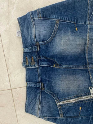 Blå jeanskjol från Monica Carina - Säljer en blå jeanskjol från Monica Carina med coola detaljer som dragkedja framtill, fickor både fram och bak samt dekorativa sömmar. Kjolen har bälteshällor och knappar i midjan för extra stilpoäng. Perfekt för dig som gillar denim och streetstyle.