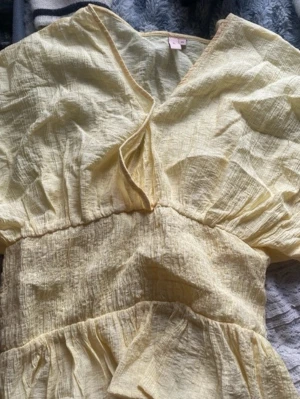 Ljusgul omlottopp med volang - Söt ljusgul omlottopp med v-ringning och volangkant nedtill. Toppen har korta, vida ärmar och smockad midja som ger en feminin siluett. Tillverkad i ett lätt, krinklat bomullstyg som passar perfekt till varmare dagar.