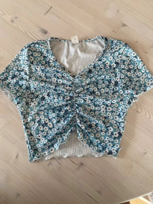 Blommig croppad topp från SHEIN - Söt croppad topp från SHEIN med blått och vitt blommönster. Toppen har v-ringning, korta ärmar och snygg rynkning framtill. Tillverkad i ribbad polyester som ger en stretchig och bekväm känsla. Perfekt för dig som gillar trendiga och färgglada plagg. Köparen står för frakt.
