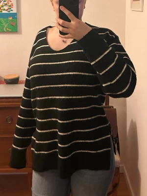 Svart stickad tröja med vita ränder - Snygg svart stickad tröja med vita horisontella ränder och V-ringning. Tröjan har ribbstickade muddar vid ärmslut och nederkant. Perfekt för dig som gillar klassiska mönster och enkel stil. Har nopprar 