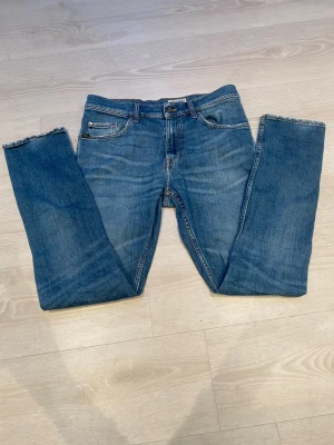 Blå jeans Tiger of Sweden raka ben - Säljer ett par klassiska blå jeans från Tiger of Sweden med raka ben och fem fickor. Jeansen har en snygg tvätt och detaljer i kontrastsömmar samt en läderpatch med logga bak i midjan. Perfekta till en avslappnad stil.