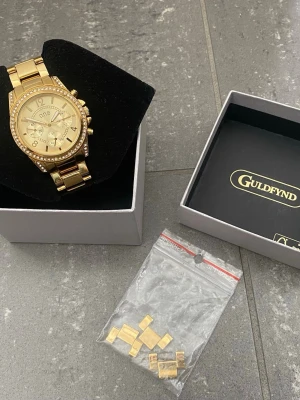 Guldig klocka från Guldfynd med stenar - Snygg guldig klocka från O.N.E med glittrande stenar runt urtavlan och länkarmband i metall. Klockan har flera små urtavlor och kommer i originalask från Guldfynd. Extra länkar till armbandet medföljer. Snabb Leverans!