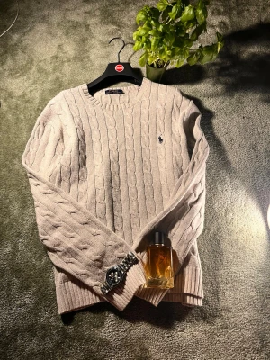 Grå kabelstickad tröja Polo Ralph Lauren - Snygg beige kabelstickad tröja från Polo Ralph Lauren med klassisk rund halsringning och den ikoniska broderade loggan på bröstet. Tröjan har långa ärmar och ribbade muddar vid ärmslut och nederkant. Perfekt för lager på lager och stilren vardagslook.