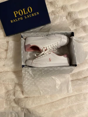 Vita Polo Ralph Lauren sneakers - Snygga vita sneakers från Polo Ralph Lauren med rosa detaljer på häl och insida. Klassisk låg modell med vita snören och diskret logga i rosa på sidan. Tillverkade i slätt läderliknande material för en clean och fräsch look.