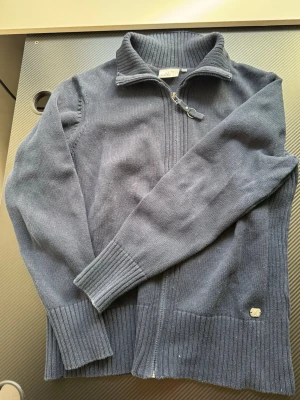 Mörkblå stickad ziptröja Isolde - Mörkblå stickad tröja från Isolde med dragkedja framtill och hög krage. Tröjan har ribbstickade muddar vid ärmslut och nederkant samt en liten metallbricka nertill. Perfekt för lager på lager och enkel att matcha.