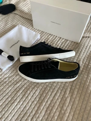 Common Projects Achilles Low Black suede sneakers - Snygga svarta sneakers från Common Projects i mocka med klassisk vit sula och guldfärgad numrering på sidan. Lågt skaft och minimalistisk design som passar till det mesta. Skorna har svarta skosnören och en stilren look.