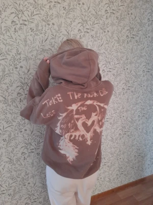 Brun hoodie med tryck och text - Cool brun hoodie med stor huva och unikt blekt tryck på ryggen. Texten 'Take the risk or lose the chance' och ett hjärta med vingar ger en edgy vibe. Perfekt för dig som gillar streetwear och vill sticka ut. Mjuk bomullskänsla och avslappnad passform.