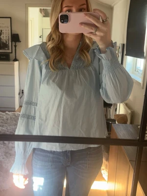 Ljusblå volangblus med lång ärm - Trendig ljusblå blus med volangdetaljer vid axlarna och smockade muddar vid ärmslut. Blusen har dekorativa sömmar och en avslappnad passform. Från Only, knappt använd