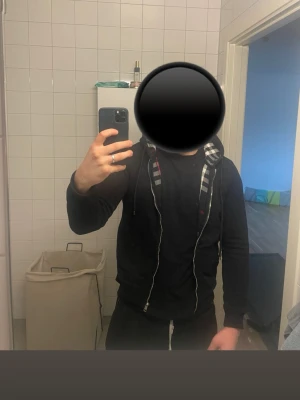 Svart hoodie med dragkedja - Säljer en svart hoodie med dragkedja och huva. Insidan av huvan har ett rutigt mönster i svart, vitt och rött. Hoodien har snören i huvan och fickor framtill. Perfekt för en avslappnad och stilren look.