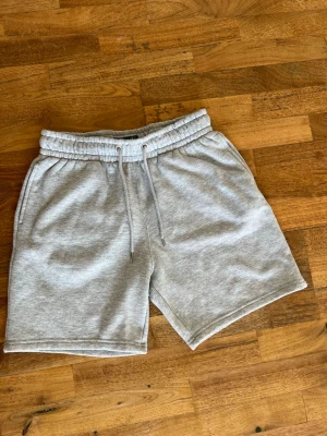 Grå sweatshorts med snörning - Säljer ett par ljusgrå sweatshorts med elastisk midja och snörning. Shortsen har två sidofickor fram och en bakficka. Perfekta för chill dagar eller träning. Mjukt material som känns skönt mot huden.