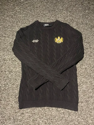 Svart kabelstickad tröja Morris - Kabelstickad svart tröja från Morris med rund halsringning. Tröjan har broderad text på bröstet och ett gult emblem med krona och lejon. Ribbstickade muddar vid ärmslut och nederkant. Perfekt för dig som gillar klassisk och stilren design. Mörkblå, storlek L men passar M