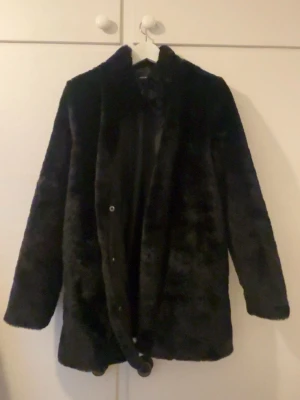 Svart fuskpäls kappa från Vero Moda - Snygg svart kappa i fuskpäls från Vero Moda. Rak modell med krage och knäppning framtill. Mjuk och fluffig känsla, perfekt för att höja din outfit under kyliga dagar. Fodrad insida för extra komfort.