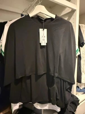 Svart cape topp i polyester - Säljer en svart cape topp från Monki i mjukt polyester-material. Toppen har en lös passform med lager på lager-effekt och rund halsringning. Perfekt för dig som gillar en unik och flowy stil.