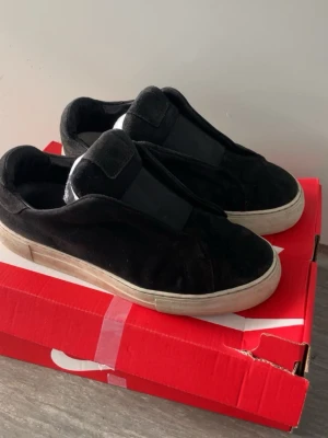 Svarta slip-on sneakers i mocka - Stilrena svarta slip-on sneakers med ovandel i mjuk mocka och breda elastiska band över vristen. Skorna har en vit, något tjockare sula och minimalistisk design utan synliga snören. Perfekta för en clean och avslappnad look.