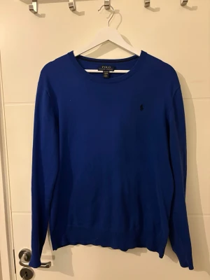 Blå tröja från Polo Ralph Lauren - Klassisk blå tröja från Polo Ralph Lauren med rund halsringning och diskret broderad logga på bröstet. Tröjan har långa ärmar och ribbade muddar vid ärmslut och nederkant. Perfekt att matcha med jeans eller chinos. Storleken är S men passar även en mindre M