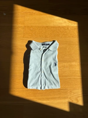 Ljusblå pikétröja från Ralph Lauren - Klassisk ljusblå pikétröja från Ralph Lauren med broderad logga på bröstet. Tröjan har krage, knappar framtill och är tillverkad i mjuk bomull. Perfekt för en clean och stilren look. Står att det är L men den passar S. Den har vissa defekter. 