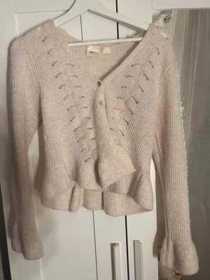 Kofta  - Söt krämvit kofta från Gina Tricot med v-ringning, dekorativa hålmönster framtill och volangkant nedtill. Koftan har långa ärmar och knäpps med tre knappar framtill. Perfekt för lager på lager och en mysig look. Aldrig använd.