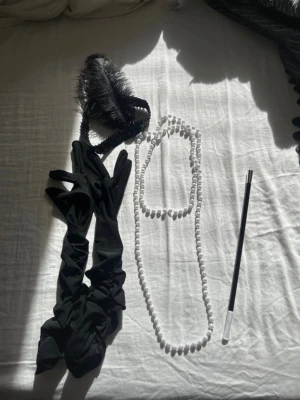 Gatsby accessoarer  - Snyggt maskeradkit i 20-talsstil med svart pannband med fjäder, långa svarta handskar, vita pärlhalsband och en svart-vit cigarettmunstycke. Perfekt för Gatsby- eller Charleston-tema och cosplay.