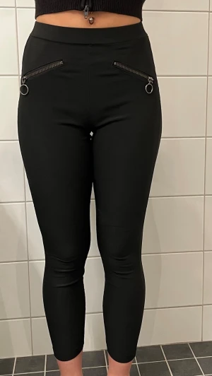 Svarta leggings med dragkedjor - Snygga svarta leggings med hög midja och två dekorativa dragkedjor framtill. Tight passform och ankellånga ben. Stretchigt material som sitter skönt och formar sig efter kroppen. Perfekta till en cool och avslappnad stil.