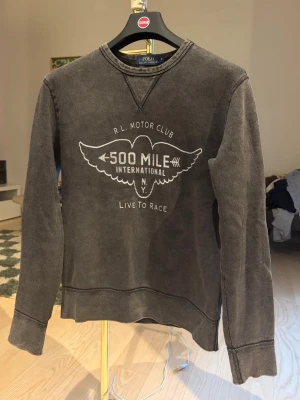 Vintage Ralph Lauren - Cool grå sweatshirt från Polo Ralph Lauren med vintage tvätt och tryck på bröstet: 'R.L. Motor Club 500 Mile International NY Live to Race'. Tröjan har rund halsringning, ribbade muddar och avslappnad passform. Perfekt för en avslappnad streetwear-look.