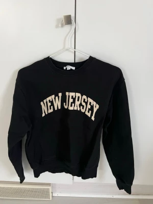 Svart sweatshirt Subdued New Jersey - Cool svart sweatshirt från Subdued med texten 'NEW JERSEY' i beige på bröstet. Klassisk rund halsringning och ribbade muddar vid ärmslut och nederkant. Perfekt för en avslappnad streetstyle-look.