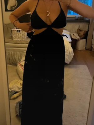 Svart långklänning med cut-outs - Snygg svart långklänning med tunna axelband och trendiga cut-outs vid midjan. Klänningen har en omlottliknande topp och är ärmlös, vilket ger en modern och stilren look. Perfekt för sommarkvällar eller när du vill sticka ut lite extra.