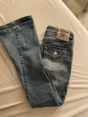 True Religion ljusblå jeans med glitter fickor  - Säljer ett par ljusblå jeans från True Religion i boutcut med snygga detaljer av glitter på bakfickorna. Jeansen har klassisk femficksdesign och en patch med logga bak i midjan. Materialet är denim i bomull och de har en cool, avslappnad vibe.
