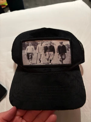 Svart truckerkeps med vintage-tryck - Säljer en svart truckerkeps med mesh på sidorna och ett stort vintage-inspirerat tryck framtill som visar fyra personer i gammaldags kläder. Kepsen har böjd skärm och justerbar baksida. Perfekt för dig som gillar retrostil och unika accessoarer.