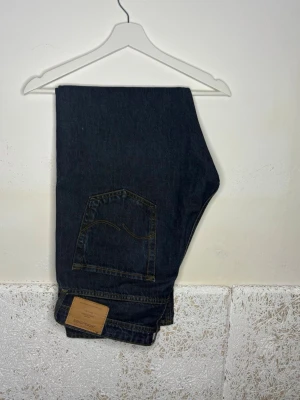 Mörkblå jeans från jack & jones - Säljer ett par klassiska mörkblå jeans med raka ben och kontrastsömmar. Jeansen har en bakficka med dekorativ söm och en läderpatch med logga vid linningen. Perfekta för en clean och tidlös look.