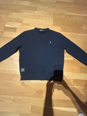 Mörkblå sweatshirt från Morris - Snygg mörkblå sweatshirt från Morris med rund halsringning och diskret logga broderad på bröstet. Tröjan har långa ärmar och en patch nertill vid midjan. Perfekt för en clean och stilren look.