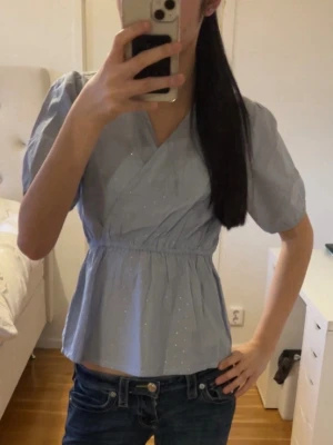 Blå omlottopp från Sofie schnoor - Söt ljusblå omlottopp från soffor schnoor med korta puffärmar och små vita prickar. Toppen har resår i midjan för en smickrande passform och v-ringad hals. Perfekt till jeans eller kjol för en fräsch look.