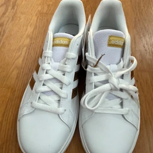 Vita Adidas sneakers med gulddetaljer - Vita Adidas Grand court med guld stripes, helt oanvända. Stl 36. Saknar kartong Nypris 900kr Intressekoll!