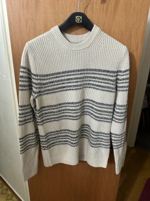 Beige stickad tröja med svarta ränder - Snygg beige stickad tröja med svarta horisontella ränder. Tröjan har rund halsringning och långa ärmar. Grovstickad struktur ger en avslappnad vibe och passar perfekt till jeans eller kjol. Materialet känns mjukt och bekvämt.