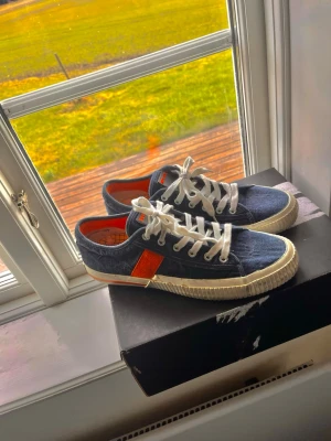 Mörkblå Replay skor med orange detalj - Snygga mörkblå Replay skor med vit sula och vita skosnören. Skorna har en orange detalj på sidan och coolt mönstrad innersula i orange och svart. Ovandelen är i tyg och modellen är klassiskt låg. Perfekta för dig som gillar retrostil och streetwear.