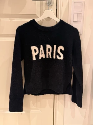 Mörkblå stickad tröja med PARIS-tryck - Cool mörkblå stickad tröja från DIVIDED med vit text 'PARIS' framtill. Tröjan har rund hals, långa ärmar och en något kortare, rak passform. Perfekt för dig som gillar streetstyle och vill ha en trendig look. Bara testat den på så den har aldrig använts. Köpt på hm!!