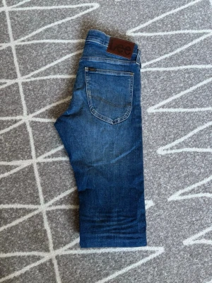 Blå slim fit jeans från Lee - Snygga blå slim fit jeans från Lee med klassisk femficksmodell och läderpatch bak i midjan. Jeansen har en lätt tvättad look och raka ben. Perfekta till vardagsstilen och tillverkade i mjuk denim för skön känsla. Storlek W27 L32. Hör av er vid frågor eller lägg ett prisförslag!