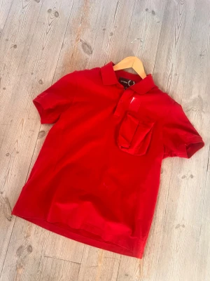 Röd pikétröja Raf Simons x Fred Perry - Säljer en röd pikétröja från Raf Simons x Fred Perry med klassisk krage, korta ärmar och en unik ficka med lock på bröstet. Tillverkad i bomull och har en stilren look som sticker ut. Rare.
