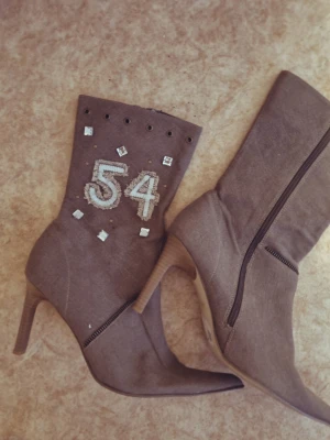 Bruna Denim boots  med design  - Unika bruna stövletter med spetsig tå och hög klack. Dekorerade med siffran 54 i silver, små fyrkantiga nitar och öljett-detaljer upptill. Dragkedja på sidan för enkel påtagning. Materialet är riktigt Denim och vintage 