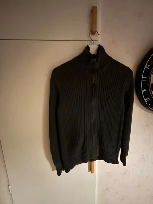 Mörkgrå ribbstickad kofta Tommy Hilfiger - Säljer en mörkgrå ribbstickad kofta från Tommy Hilfiger med dragkedja framtill och hög krage. Koftan har långa ärmar och är tillverkad i ett mjukt material som passar perfekt för kyligare dagar. Klassisk och stilren design.