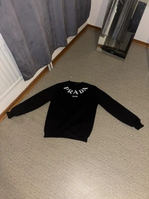 Prada tröja - Svart  tröja från Prada med vit logga och texten 'Milano' på bröstet. Tröjan har rund halsringning och långa ärmar. Enkel och stilren design som passar perfekt till dig som gillar exklusiva märken.