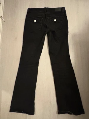 Svarta bootcut jeans med silverknappar - Snygga svarta bootcut jeans med silverfärgade knappar på bakfickorna och klassisk femficksdesign. Jeansen har låg midja och lätt utsvängda ben. Perfekta till sneakers eller boots för en cool vardagslook.