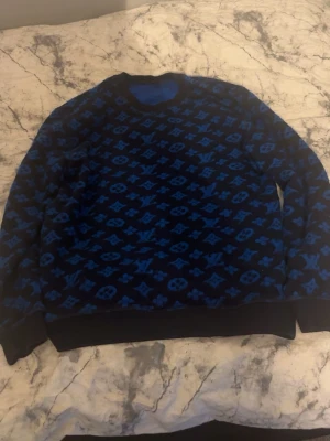 Blå Louis Vuitton monogramtröja - Säljer en blå och svart tröja från Louis Vuitton med det klassiska monogrammönstret över hela plagget. Tröjan har rund halsringning, långa ärmar och ribbade muddar vid ärmslut och nederkant. Perfekt för dig som gillar exklusiv streetwear.