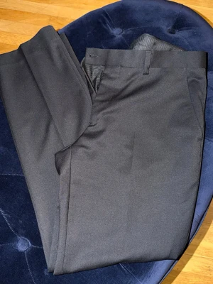 Svarta klassiska kostymbyxor - Snygga svarta kostymbyxor med rak passform och pressveck. Byxorna har bälteshällor och sidofickor, perfekta för en stilren look. Materialet ser ut att vara en slät och mjuk textil som ger en elegant känsla.