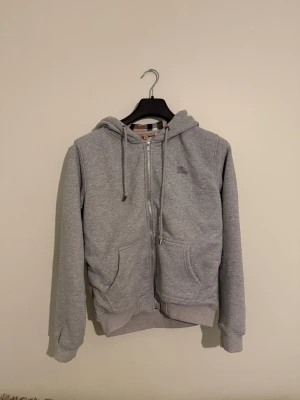 Burberry Zip Hoodie - Säljer en stilren grå hoodie från Burberry med dragkedja framtill och klassiska Burberry-ränder i luvans insida. Hoodien har två fickor, ribbade muddar och justerbar huva med snören. Perfekt för en avslappnad och trendig look.