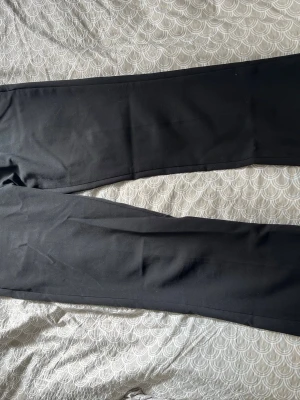 Svarta klassiska kostymbyxor - Säljer ett par svarta kostymbyxor med raka ben och pressveck. Byxorna har hällor för skärp och två bakfickor med minimalistisk design. Perfekta för en stilren look och passar till många olika outfits.