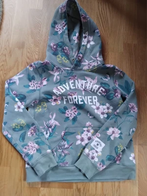 Grön hoodie med blommigt tryck - Grön hoodie från H&M med blommigt mönster i rosa, vitt och grönt. Texten 'ADVENTURE FOREVER' i vitt framtill. Mjuk och gosig känsla för en chill dag i skolan. Perfekt för dig som gillar en chill och färgglad stil.
