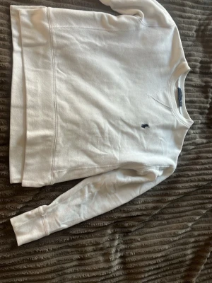 Vit sweatshirt från Polo Ralph Lauren - Klassisk vit sweatshirt från Polo Ralph Lauren med rund halsringning och den ikoniska lilla marinblå logon broderad på bröstet. Tröjan har långa ärmar och ribbade muddar vid ärmslut och nederkant. Perfekt för en clean och stilren look.
